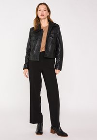 Giacca di pelle nera con due tasche frontali, abbinata a un top beige e pantaloni neri a gamba larga. Il modello indossa stivali neri alla caviglia.