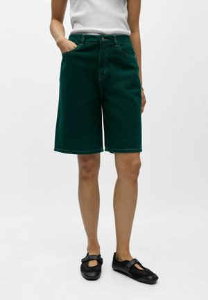 Kvinde iført mørkegrønne knælange shorts med forlommer, en hvid ærmeløs top og sorte Mary Jane-sko.