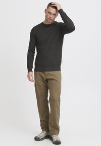 Suéter de punto acanalado gris oscuro, pantalones cargo caqui con bolsillo lateral y zapatos de senderismo grises. El modelo está de pie con la mano en la cabeza, fondo neutro.