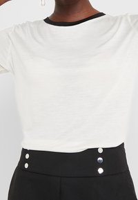 Vit t-shirt med kort ärm och svart ribbad krage, bärs tillsammans med högmidjade svarta byxor med tre metallknappar i midjan.