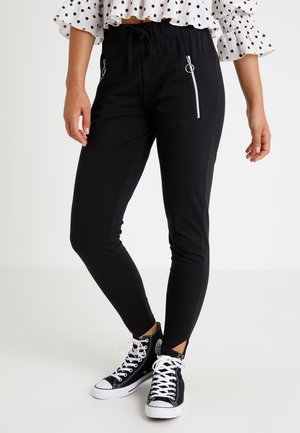 Femme portant un pantalon noir ajusté avec poches à fermeture éclair, associé à des baskets montantes noires et un crop top à volants blanc à pois.