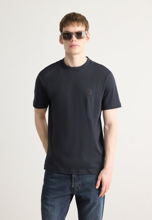 Dark grey cotton t-shirt with short sleeves and a crew neckline, featuring a small black logo on the chest. Worn with denim jeans.