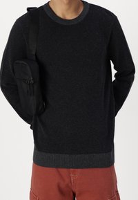 Uomo che indossa un maglione nero texturizzato, pantaloni rossi, con una piccola borsa a tracolla nera sulla spalla sinistra, mani dietro la schiena su sfondo bianco.