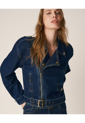 Frau mit langen Haaren, die eine dunkelblaue Jeansjacke mit Reißverschluss, goldenen Knöpfen und passende Jeanshose vor einem schlichten Hintergrund trägt.