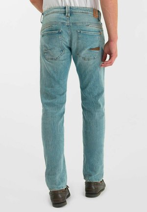 Man draagt lichtblauwe spijkerbroek en donkerbruine schoenen, staat met de rug naar de kijker toe tegen een effen witte achtergrond.