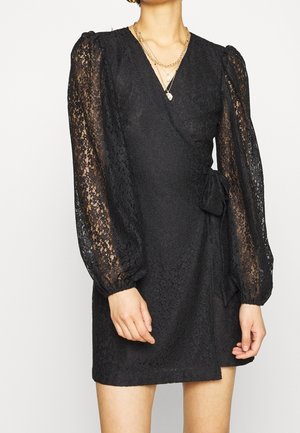 Woman wearing a black lace wrap dress with sheer long sleeves and layered gold necklaces, shown from neck to mid-thigh.