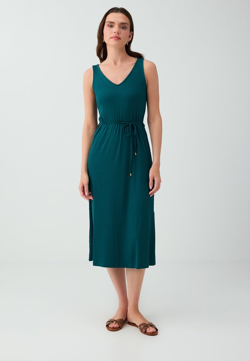 Femme debout portant une robe midi sans manches bleu sarcelle avec une ceinture à la taille et des sandales plates marron sur un fond clair uni.
