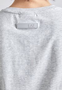 Tricou gri din bumbac cu guler rotund și mâneci scurte. Are o etichetă rectangulară gri cu marca "EA7" la gulerul din spate.