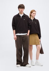 Νεαρός άνδρας και γυναίκα που φοράνε μαύρα πουλόβερ με φερμουάρ. Ο άνδρας φορά καφέ παντελόνι, η γυναίκα κίτρινη καρό φούστα και λευκά αθλητικά παπούτσια, κρατώντας καφέ τσάντα.