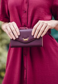 Burgundy wallet with textured finish and gold zigzag stitching on the front, held between hands, paired with a matching burgundy dress.
