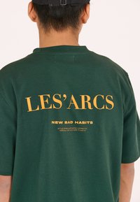 Vihreä puuvillapaita, jossa on suuri keltainen teksti "LES'ARCS" ja pienempi teksti "NEW BAD HABITS" takana. Lyhyet hihat ja rento istuvuus.
