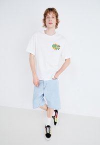 Hvit bomull t-skjorte med grafisk trykk på venstre bryst, blå denim-shorts og svarte joggesko med rød og gul flammekdesign.