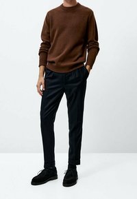 Brązowy dzianinowy sweter, czarne spodnie z zakładkami i czarne zamszowe buty. Strój charakteryzuje się nowoczesnym, czystym krojem oraz prążkowanymi mankietami.