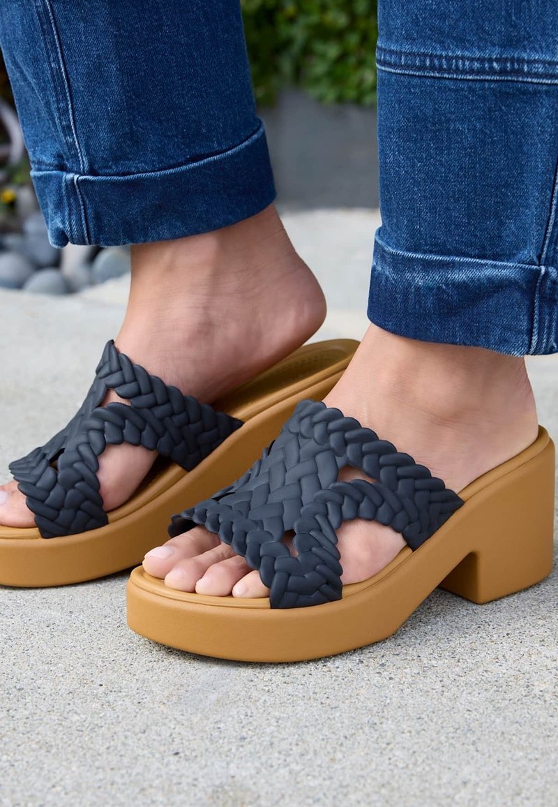 Pieds chaussés de sandales compensées couleur fauve avec de larges brides noires tressées, associées à un jean bleu retroussé, debout sur une surface en béton.