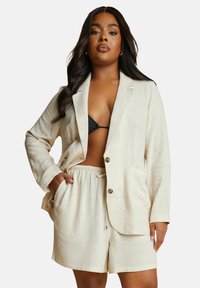 Beige linn blazer med två knappar, sidofickor och matchande dragsko-shorts. Texturerad tyg, avslappnad design, öppen frontstil.