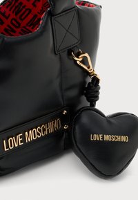 Bolsa de mão de couro preto com ferragens em tom de ouro, apresentando um chaveiro em forma de coração. Interior forrado com um padrão vermelho, logotipo em relevo na bolsa.