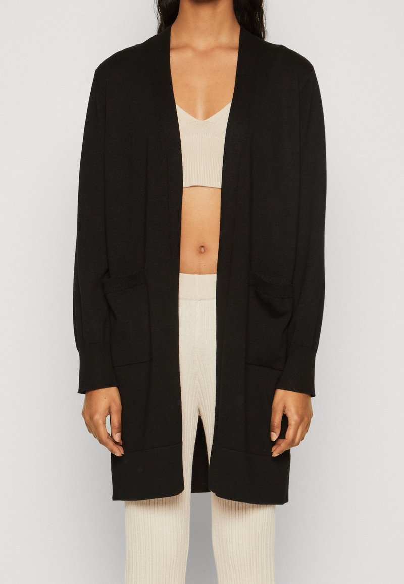 Black cardigan with long sleeves, open front, two front pockets, and ribbed detailing at cuffs and hem, worn over a beige top.