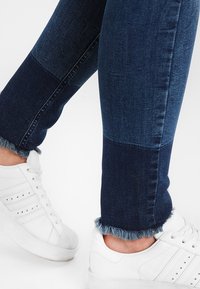 Pantalones vaqueros de denim azul oscuro con un diseño de patchwork de diferentes tonos, con dobladillos deshilachados, llevados con zapatillas blancas.