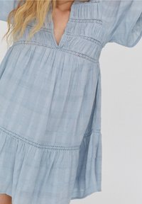 Světle modré bavlněné šaty s výstřihem do V, rozšířenými rukávy a vrstveným designem. Obsahují jemné krajkové detaily a strukturované vzory tkaniny.