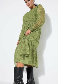 Groen jurk met patroon, lange mouwen, gerimpelde taille en asymmetrische zoom. Gecombineerd met kniehoge zwarte laarzen.