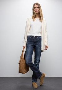Cárdigan beige, blusa blanca, jeans azul oscuro, zapatos de ante marrones y un bolso marrón. Un atuendo casual con texturas suaves y un corte relajado.