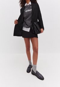 Zwarte blazer over een grijze sweatshirt met de tekst "Urban", witte blouse eronder, zwarte rok, grijze sokken en zwarte loafers. Zwarte tas.