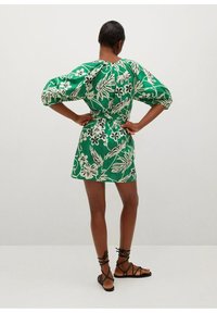 Robe courte verte à motifs floraux blancs, manches bouffantes, taille cintrée et ourlet légèrement évasé. Associée à des sandales noires à lanières.