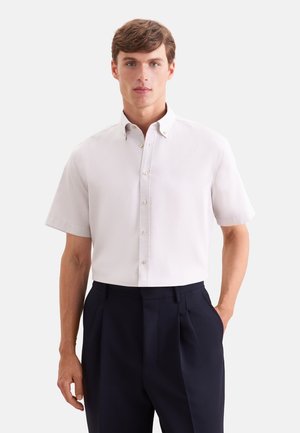 Mann trägt ein weißes kurzärmeliges Hemd mit Knöpfen, das in hoch taillierte schwarze plissierte Hose gesteckt ist, steht vor einfachem Hintergrund.