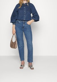 Jeansskjorta med puffärmar, knappstängning framtill, ljusblå jeans, metalliska blockklack-sandaler och en texturerad handväska i rosa ormskinnsmönster.