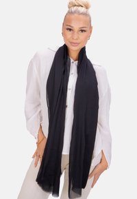 Écharpe noire légère qui s'enroule doucement, fabriquée en tissu texturé. Portée sur une chemise blanche à boutons, elle complète un pantalon de teintes neutres.