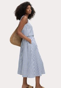 Abito midi a strisce blu e bianche con top senza maniche, tasche laterali e gonna svasata, abbinato a una borsa a tracolla intrecciata.