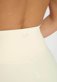 Leggings taille haute en tissu côtelé crème clair avec une texture lisse et extensible. Présente un logo S/B discret au niveau de la taille.
