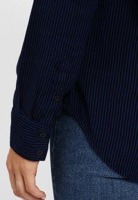 Granatowa koszula w paski z poziomymi liniami, tkanina o fakturze, z zapięciem na guziki przy mankietach. Noszona z dopasowanymi dżinsami.