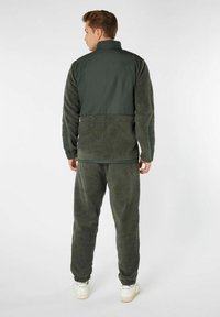 Tuta in pile di colore verde scuro, composto da una giacca con zip e accenti trapuntati e pantaloni abbinati con polsini a coste. Tessuto morbido e testurizzato.