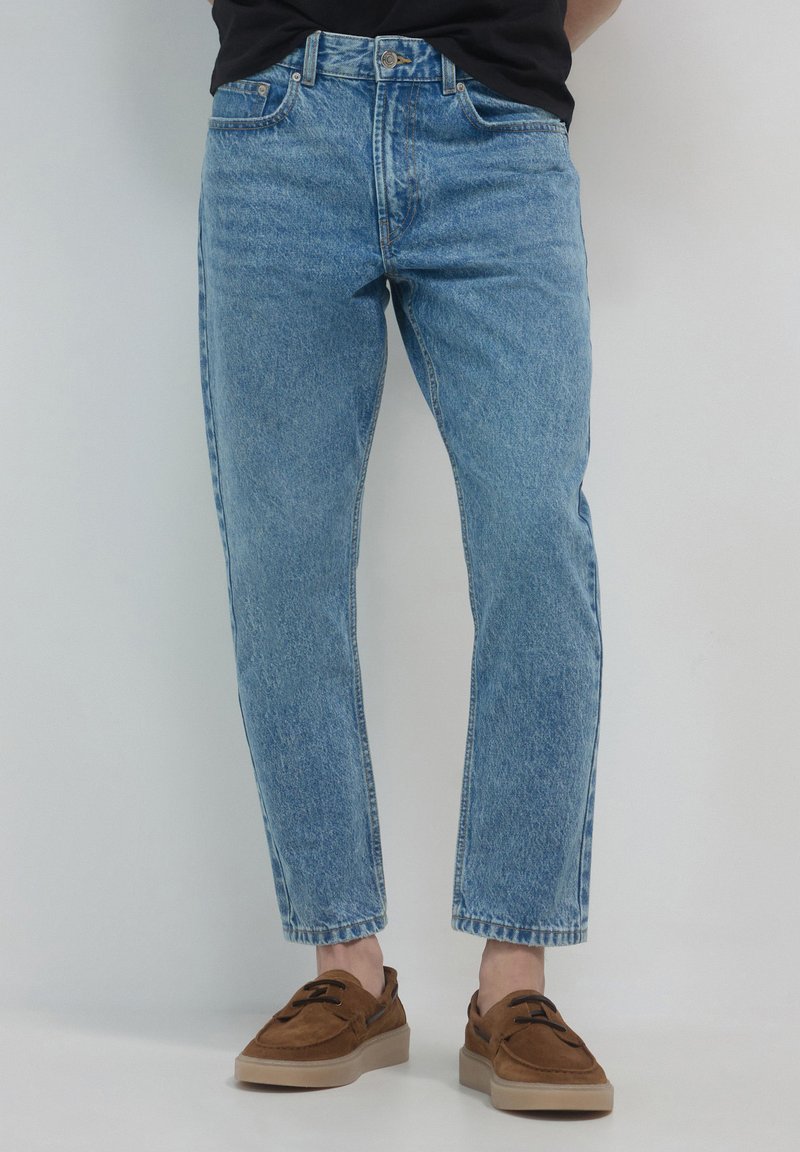 Jeans en denim bleu clair avec une coupe décontractée, texture délavée, design à cinq poches et longueur courte, associés à des chaussures marron.