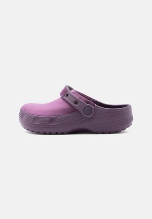 Lilla slip-on træsko med tekstureret sål og justerbar hælrem set i profil mod en ensartet lys baggrund.