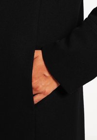 Svart yllekappa med en sidoficka, närbild som visar en hand som vilar inuti fickan. Har en mjuk textur och en strukturerad design.