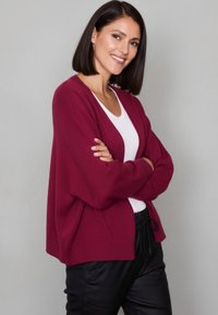 Burgundy cardigan with a loose fit, ribbed cuffs and hem, styled over a white shirt. The fabric appears soft and textured.