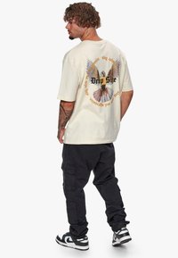T-shirt crème surdimensionné avec un graphisme d'oiseau et du texte au dos, associé à un pantalon cargo noir et des baskets noir et blanc.