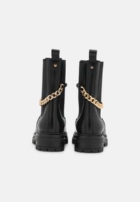 Czarne skórzane botki z chamskimi podeszwami i złotym łańcuchem z tyłu, pokazane z tyłu na białym tle.