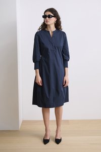 Tumedat sinise puuvillane kleit ümmarguse kraega, nööpidega esiosas ja puhvitud varrukatega. Stiilitud mustade teravate varvastega kingade ja prillidega.