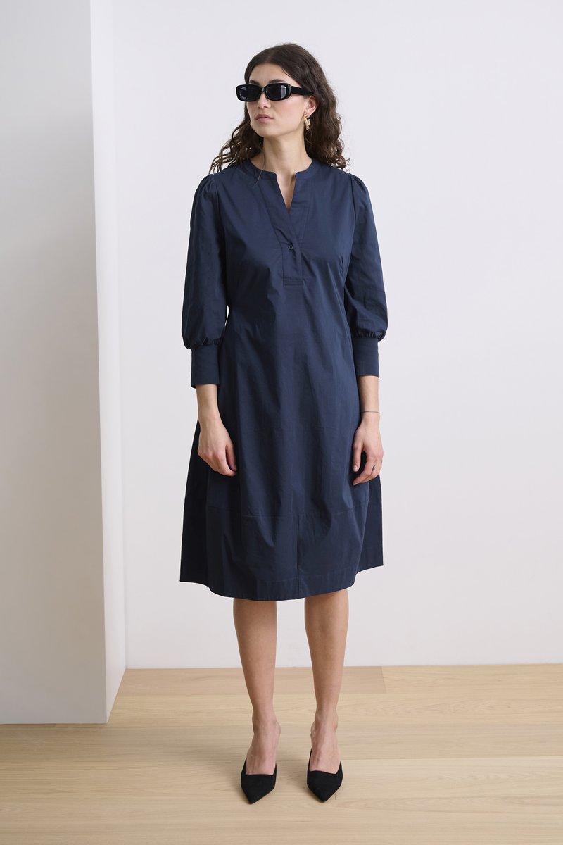 Tumedat sinise puuvillane kleit ümmarguse kraega, nööpidega esiosas ja puhvitud varrukatega. Stiilitud mustade teravate varvastega kingade ja prillidega.