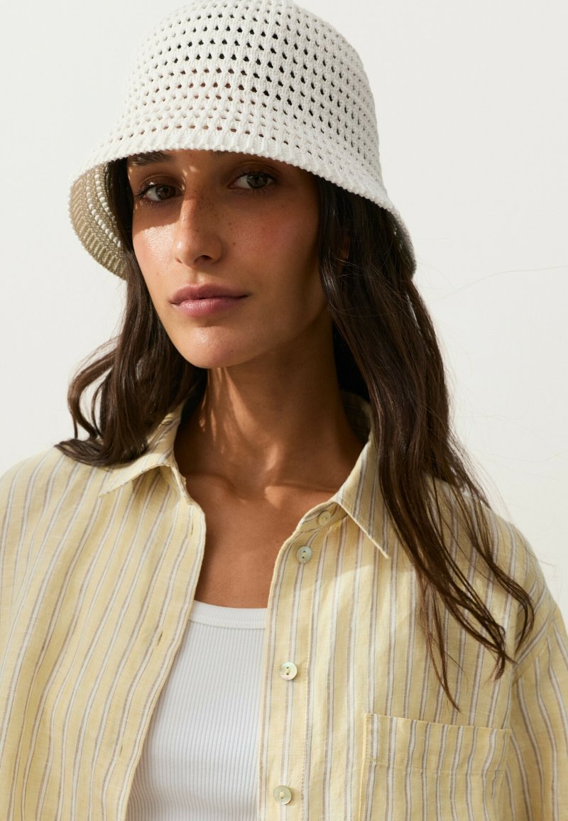 Femme aux longs cheveux foncés portant un chapeau bob blanc en crochet, une chemise rayée jaune à boutons et un débardeur blanc côtelé, fond neutre.
