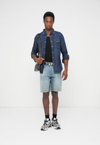 Chemise en denim, t-shirt noir, short en denim bleu clair, et chaussettes noires avec des baskets blanches. Sac bandoulière avec un motif.