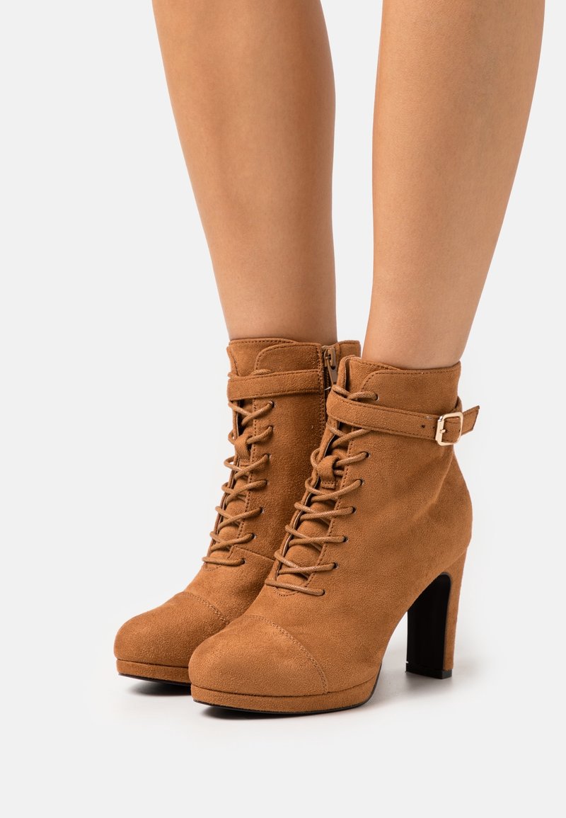Bottines en daim marron avec talons carrés hauts, lacets à l'avant, fermeture éclair latérale et une sangle décorative avec boucle autour de la cheville.