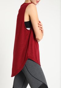 Punainen hihaton toppi, jossa on kaareva helma, rento istuvuus ja sekoitus erilaisia pintoja, yhdistettynä harmaisiin legginsseihin, joissa on erottuvat saumat.