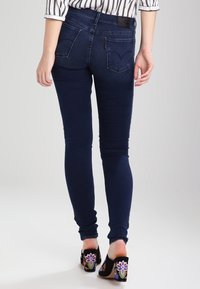 Mujer con jeans ajustados azul oscuro y tacones cuadrados bordados en negro, de pie contra un fondo claro y uniforme.