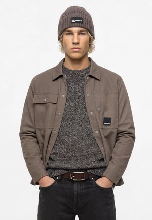 Jeune homme portant un bonnet en maille gris, une veste marron et un pull gris, debout les mains dans les poches devant un fond clair uni.