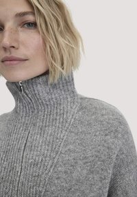 Blonde Frau mit kurzen Haaren, die einen grauen, gerippten, hochgeschlossenem Reißverschluss-Pullover trägt, aus der Nähe auf hellem Hintergrund.