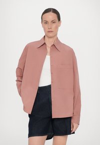 Chemise rose à manches longues, avec une poche poitrine unique et une texture de tissu lisse, associée à un short en denim foncé.